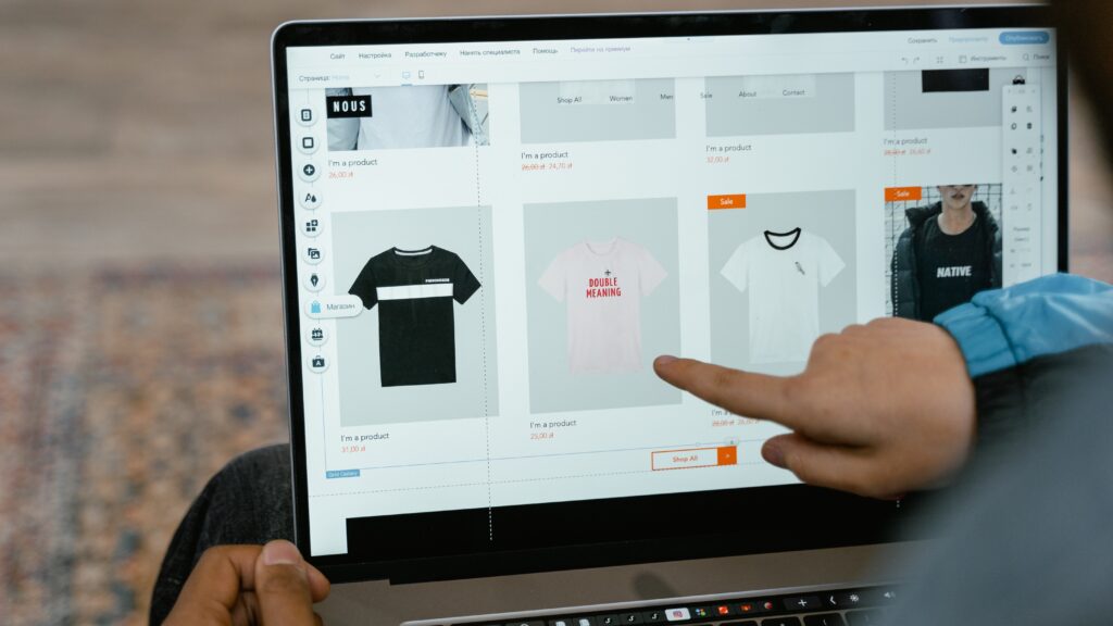 (Para todos verem). Uma pessoa branca aponta para a tela de um computador que exibe a imagem de um site de vendas online de roupas.